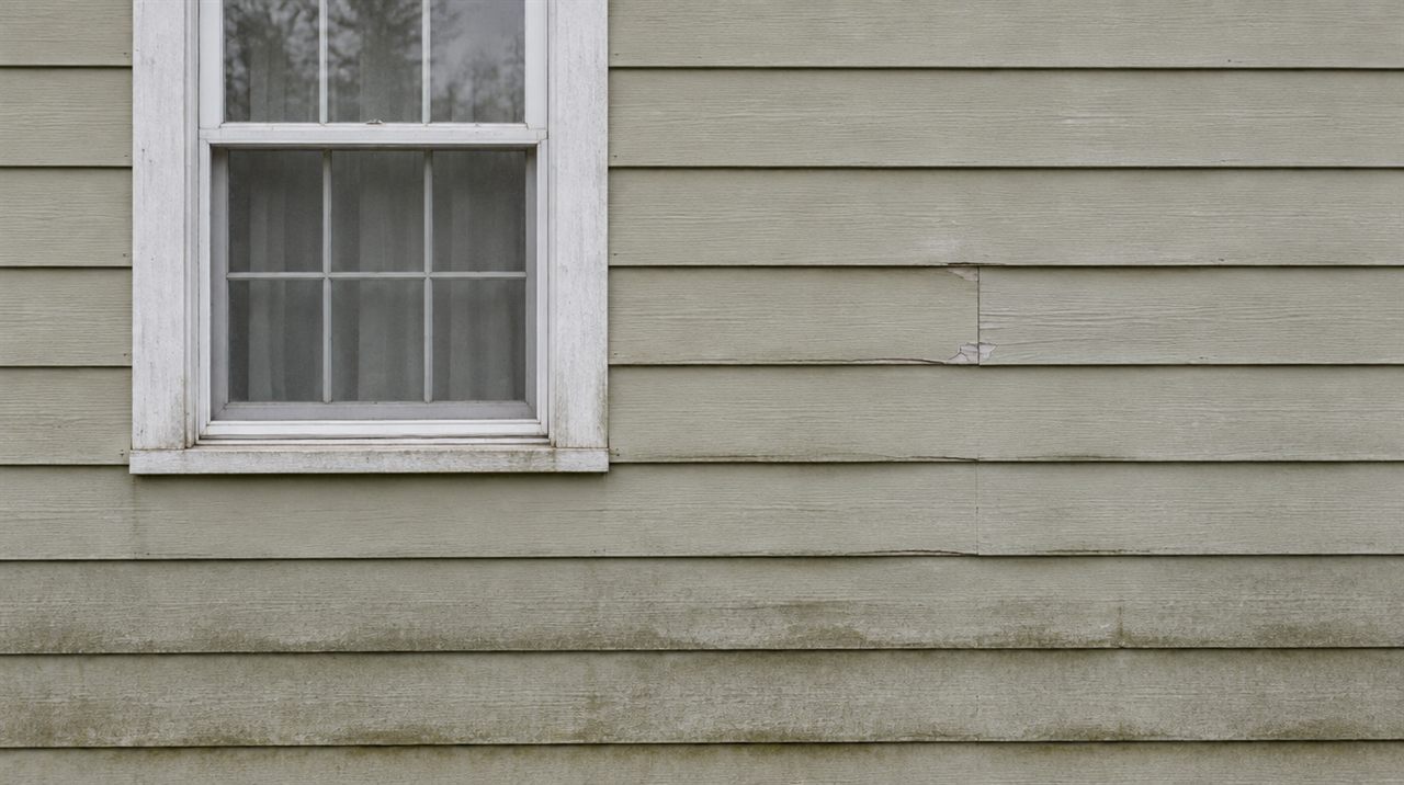 A wall before the siding replacement: sun-faded chalky paint on wood lap siding, green-gray algae grime darkening the lower courses, a split board and a patch of flaking paint, beside a window with dulled trim